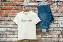 Фото бежевой футболки и синих джинсовых брюк Woodcutter на фоне грубой кирпичной стены