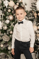 Детский фотопортрет мальчика в белой рубашке с бабочкой в динамичной позе на фоне новогодней ёлки