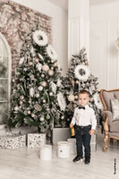 Фото малыша в полный рост в студии в белой рубашке с бабочкой на фоне новогодних ёлок