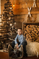 Детский новогодний фотопортрет мальчика в студии на фоне деревянной стены, деревянной елки и других декораций из дерева