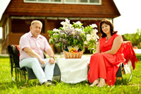 Парный семейный фотопортрет на природе (супружеская пара сидит в креслах, рядом стоит столик с букетом сирени и корзина с фруктами)