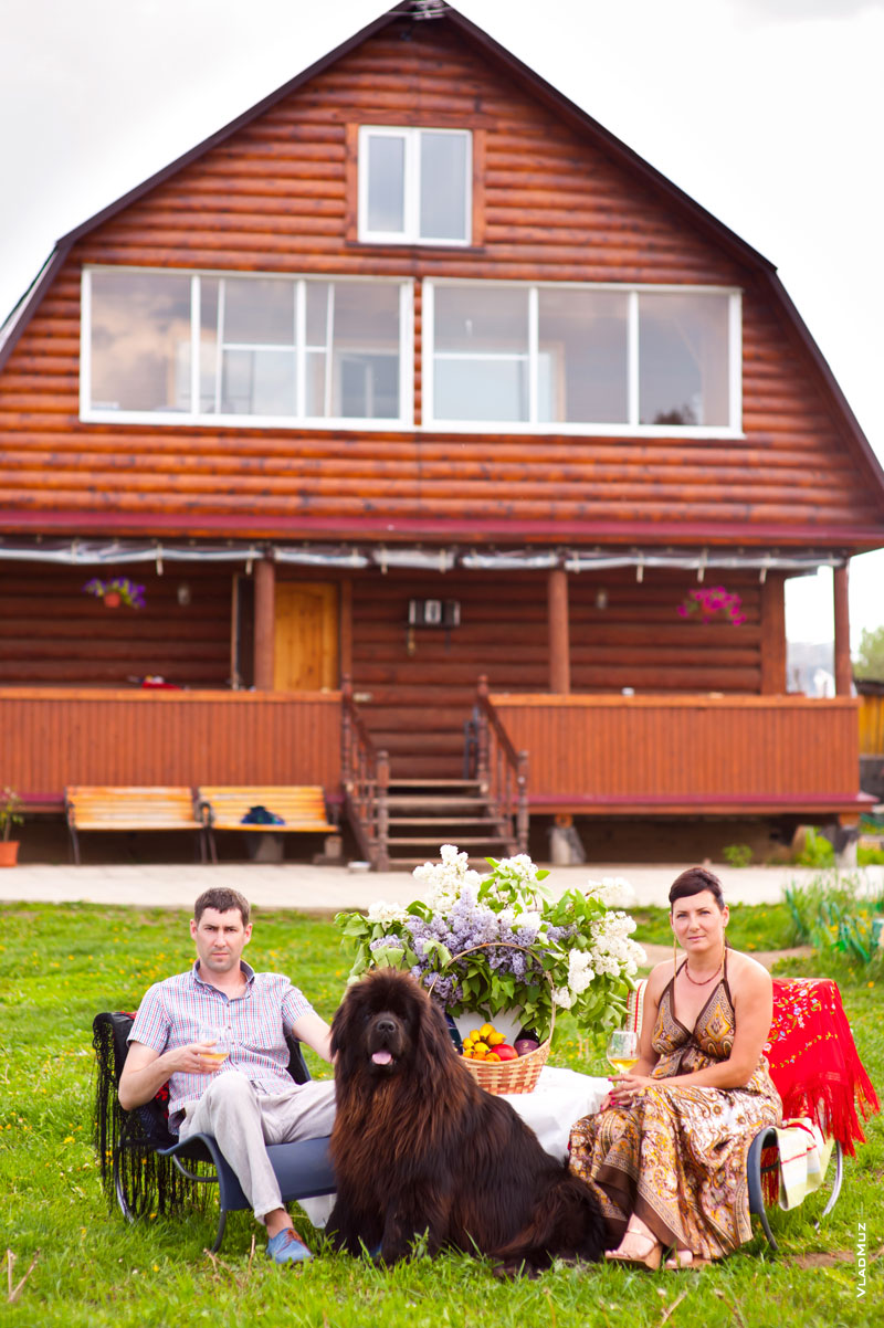 Семейное фото на природе: муж с женой с бокалами - в креслах, рядом черный ньюфаундленд, между ними - столик с букетом сирени