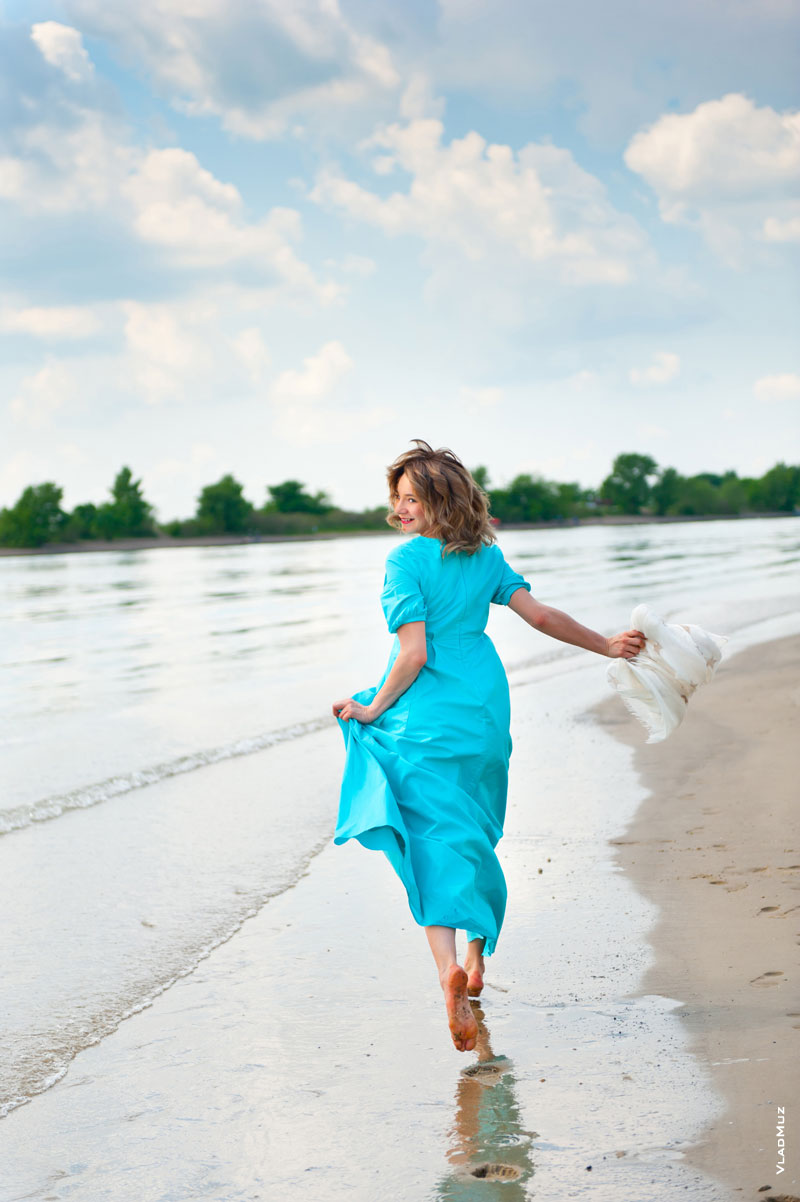 На фото девушка в бирюзовом платье бежит по песчаному берегу у кромки воды и оглядывается назад