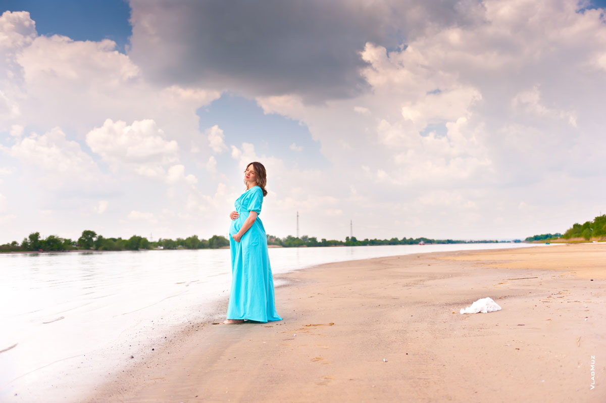 Фото беременной девушки в платье, стоящей у кромки воды на песчаном берегу