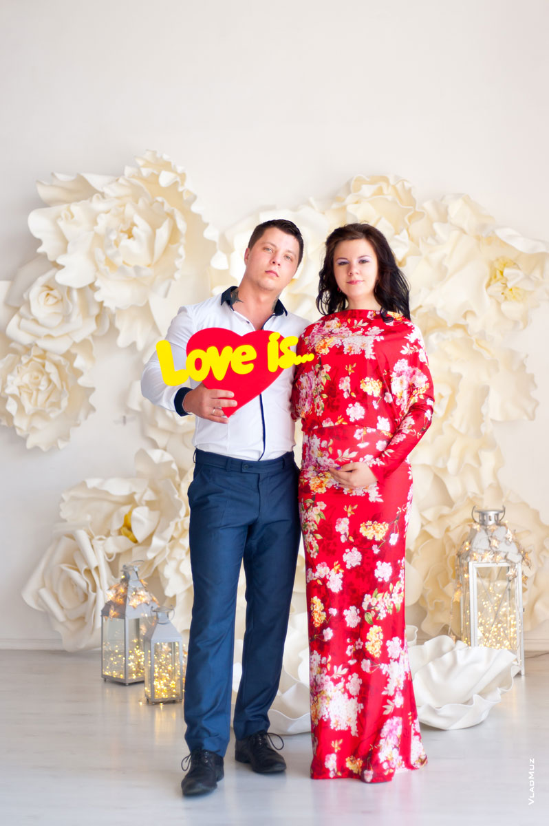 Семейное фото в полный рост: беременная женщина в красном длинном платье, рядом мужчина с буквами “Love is” на фоне сердца