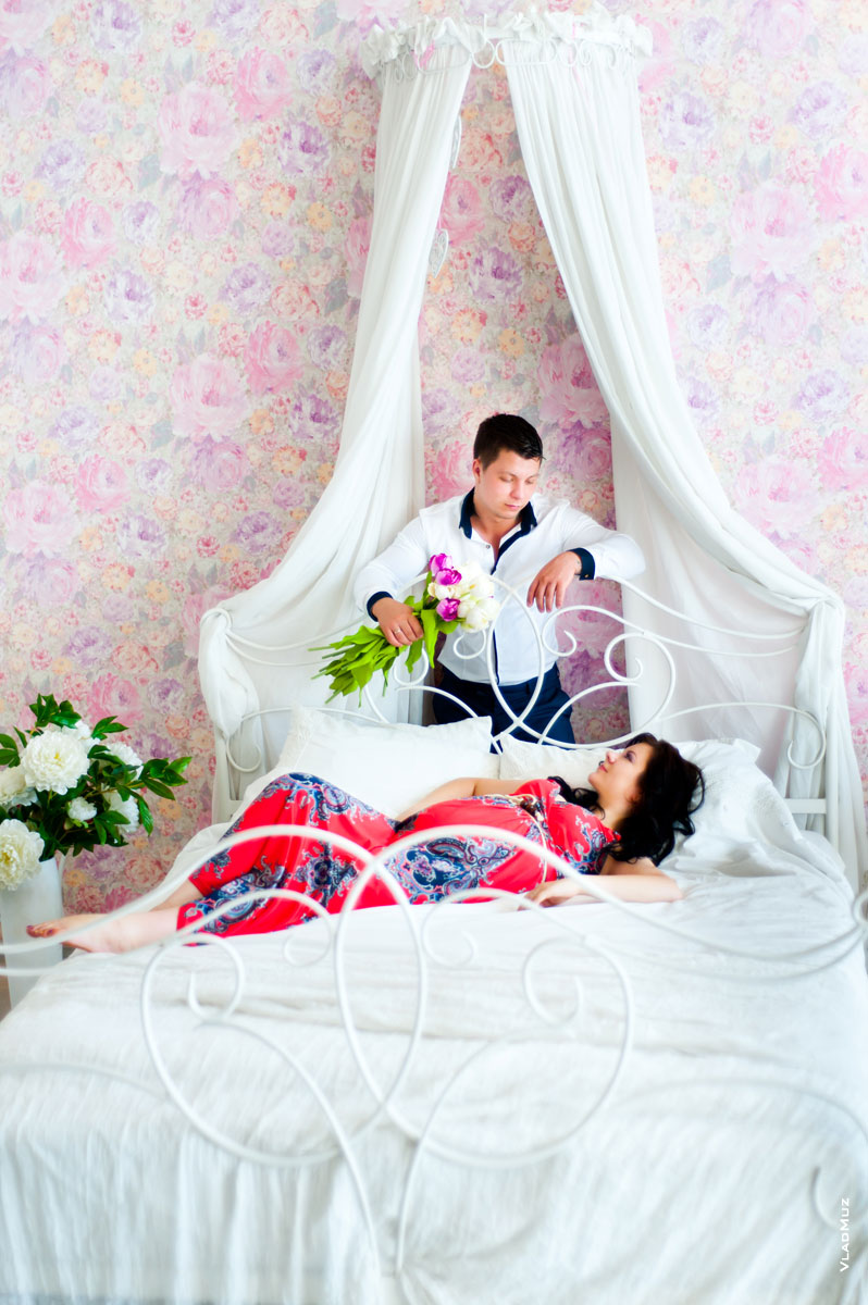 Семейное фото в спальне: на кровати беременная жена, рядом муж с цветами
