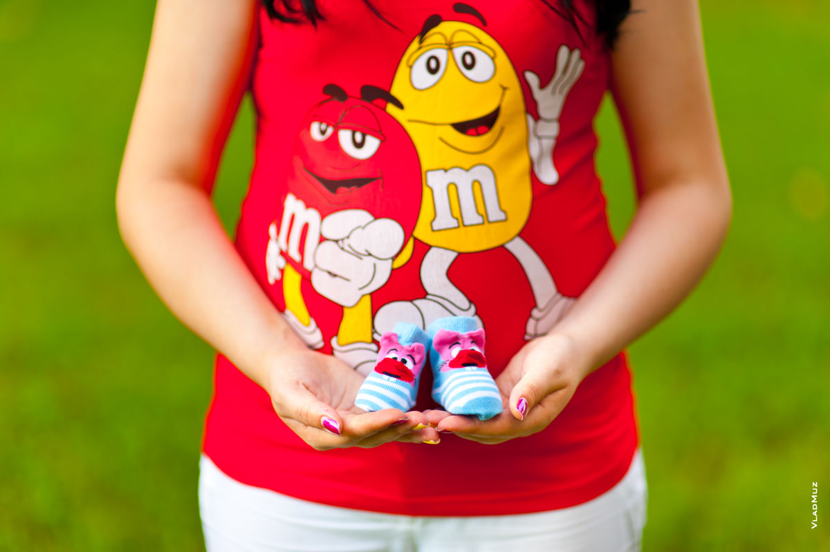 Фото беременной в красной яркой и веселой майке с голубыми пинетками в руках на фоне живота