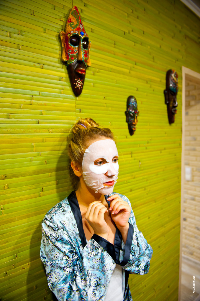 Жанровый фотопортрет девушки в косметической маске на фоне коллекции деревянных масок