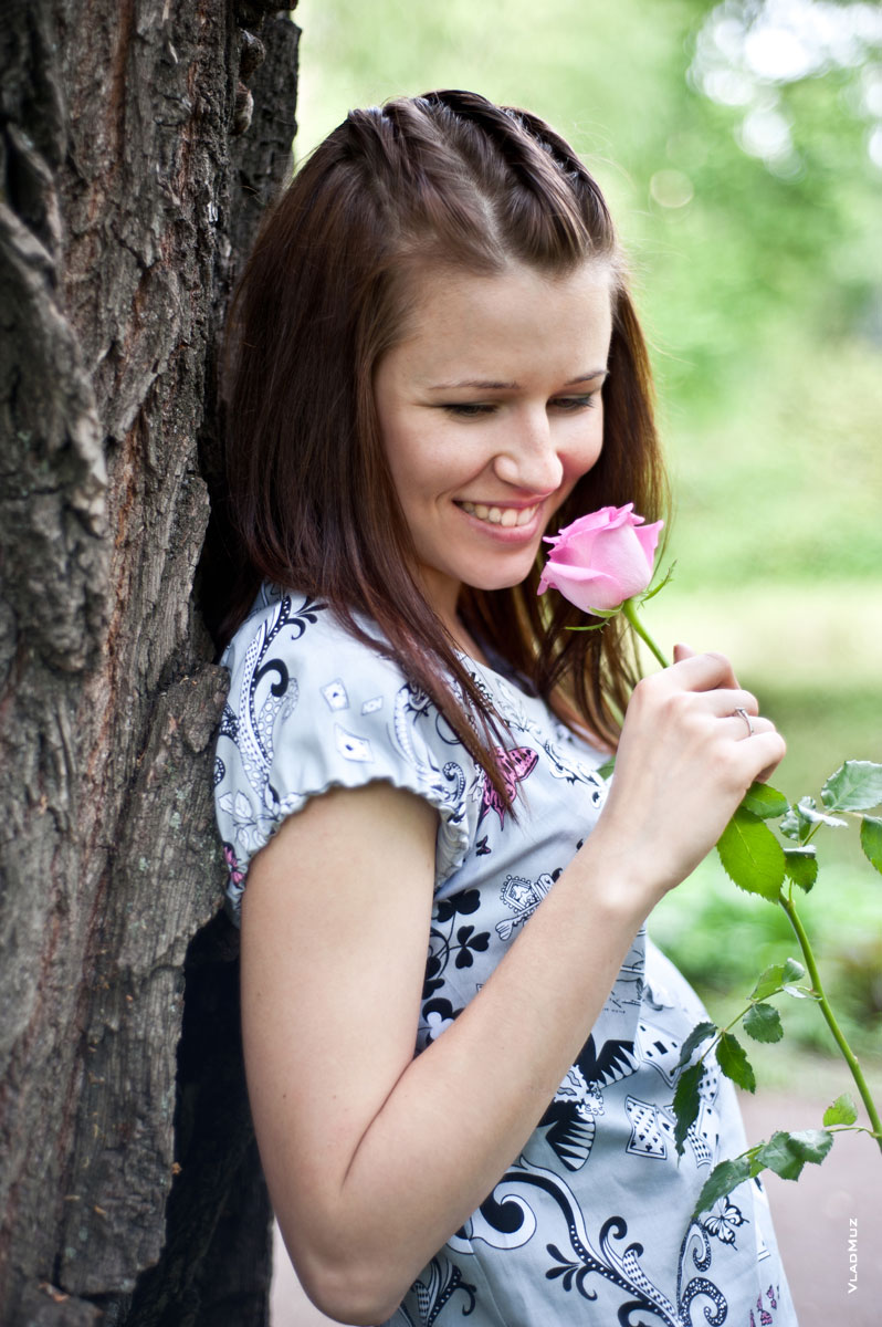 Фото улыбающейся девушки, которая стоит с розой спиной к дереву