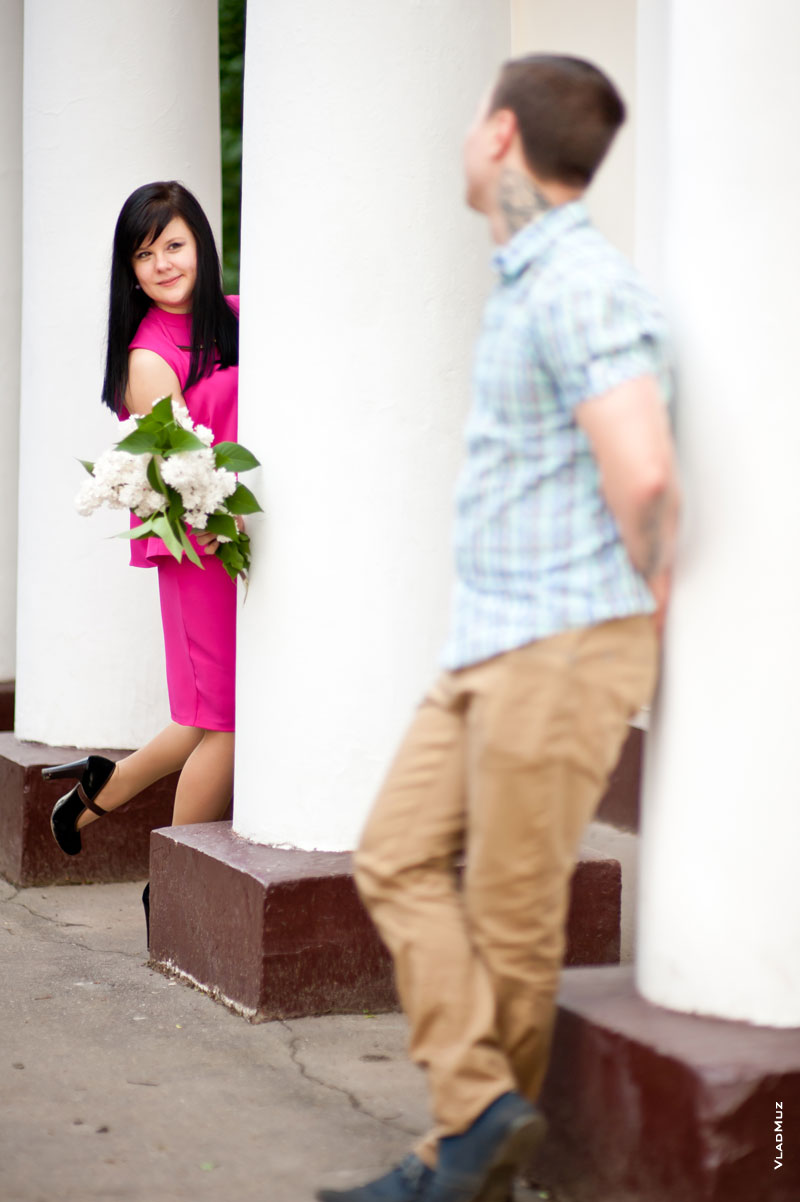 Фото мужчины у белой колонны, девушка в розовом платье с букетом выглядывает из-за колонны рядом