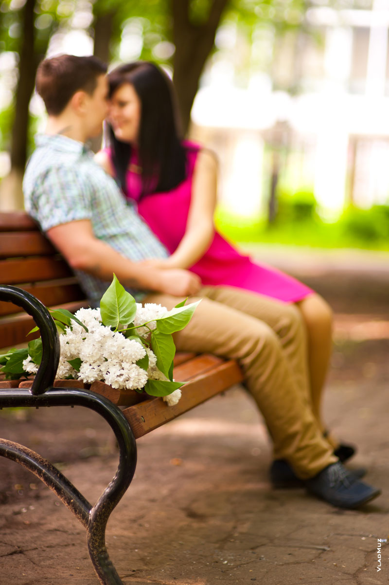 Фото букета белой сирени на скамейке в фокусе, пара влюбленных в расфокусе вдали