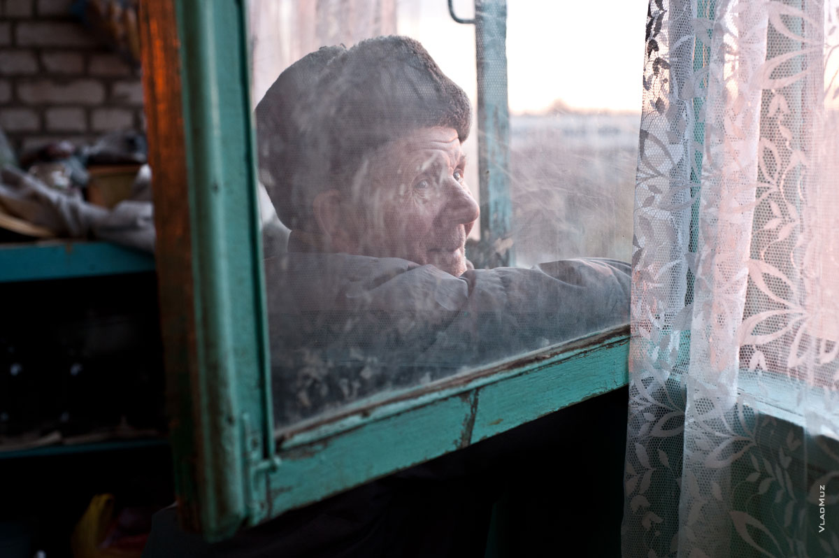 Жанровое фото сквозь грязное стекло пожилого мужчины в шапке-ушанке, смотрящего в окно