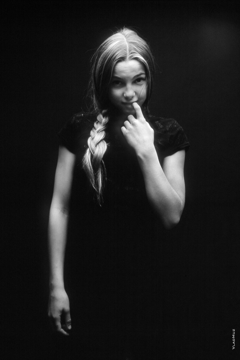 Фотопортрет смущенной девочки с пальцем во рту на черном фоне в темной тональности