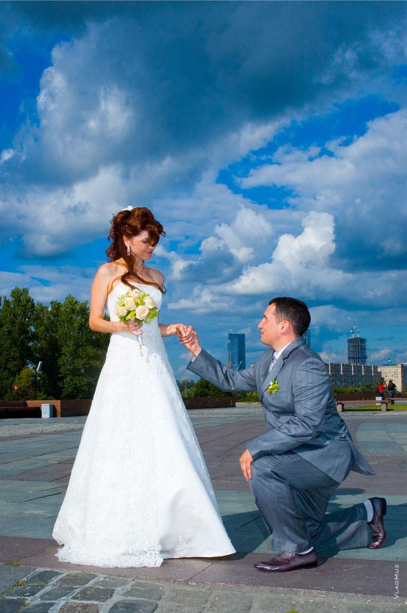 Фото невесты и жениха на колене перед ней на фоне синего неба