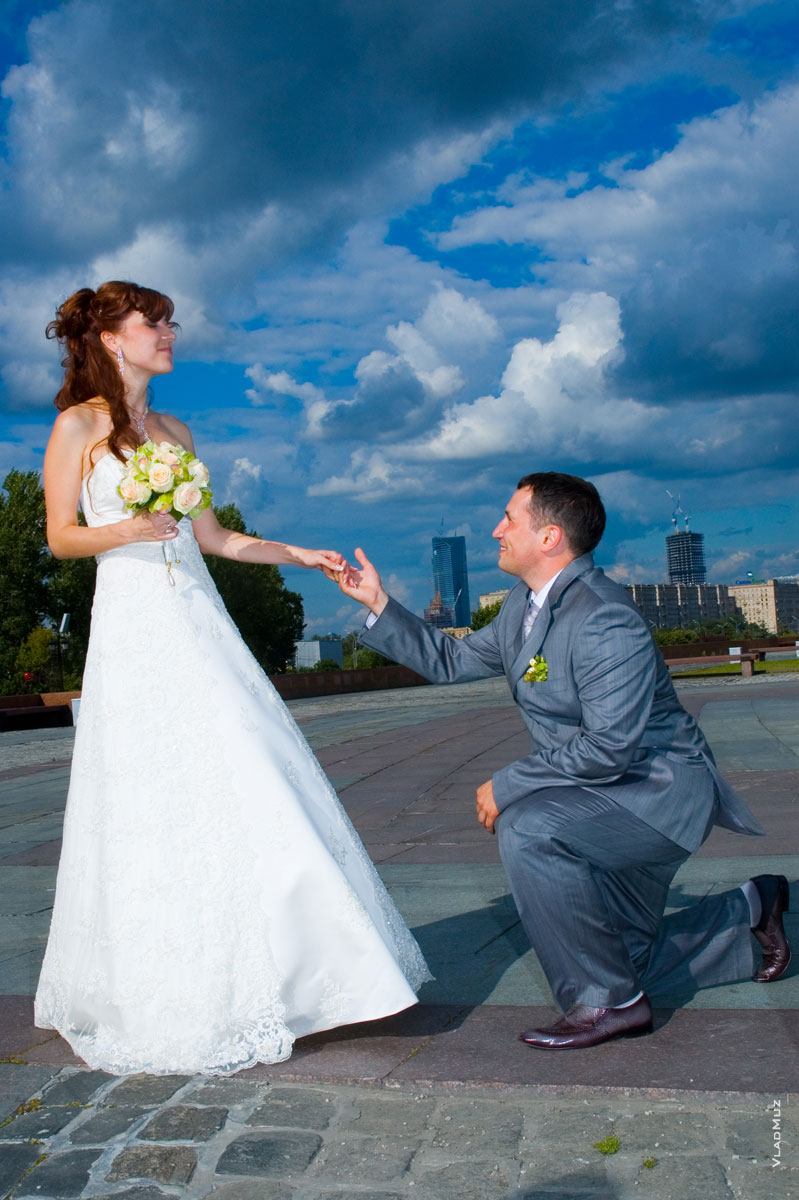Фото невесты и жениха на колене перед ней на фоне ярко-синего неба