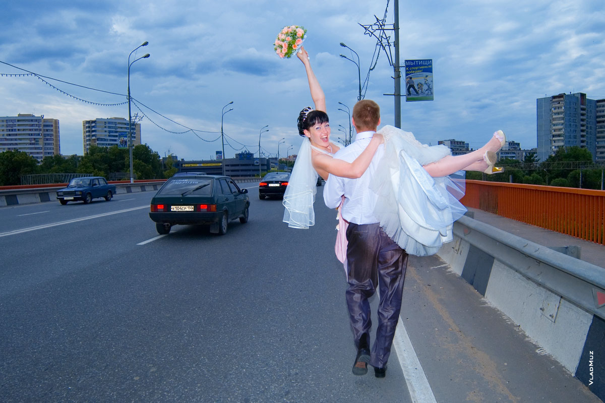 Фото жениха с невестой на руках, идущего по мосту через железную дорогу (по Олимпийскому проспекту в Мытищах)
