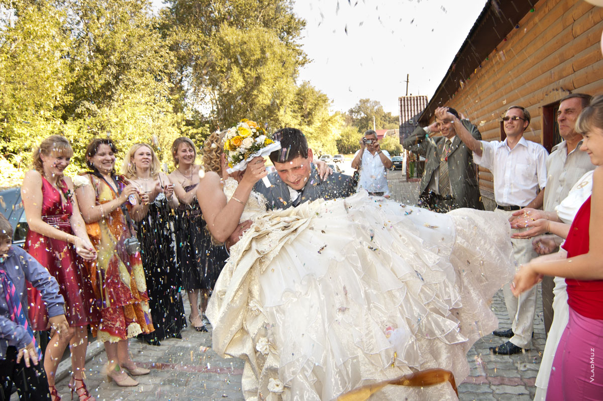 Репортажное свадебное фото: жених с невестой на руках под дождем из пшена, конфет и монет