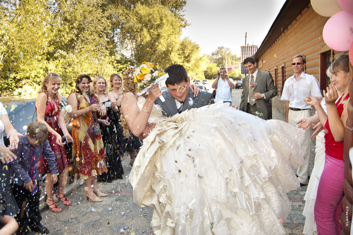 Фото жениха с невестой на руках перед рестораном под дождем из пшена, конфет и монет