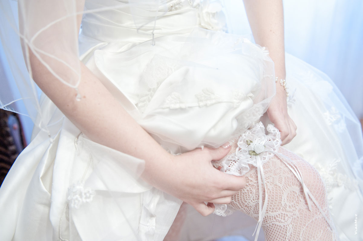 На фото руки невесты, платье и свадебная подвязка на бедре