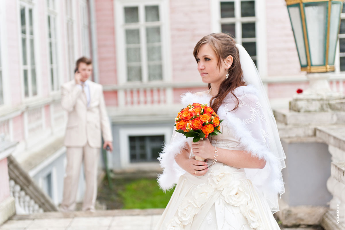 На переднем плане — невеста с букетом, вдалеке — жених говорит по телефону