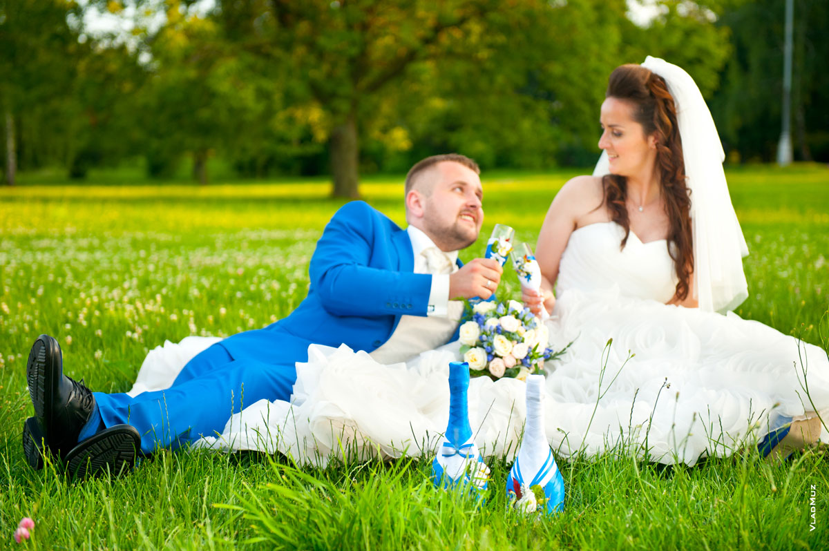 Фото на лужайке: свадебные быки на переднем плане в фокусе, на втором плане свадебная пара с бокалами шампанского