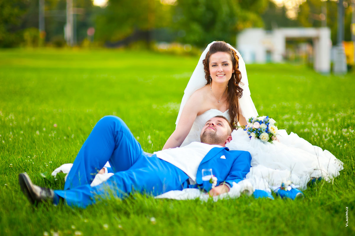 Фото лежащего на лужайке жениха с бокалом шампанского и сидящей рядом невесты
