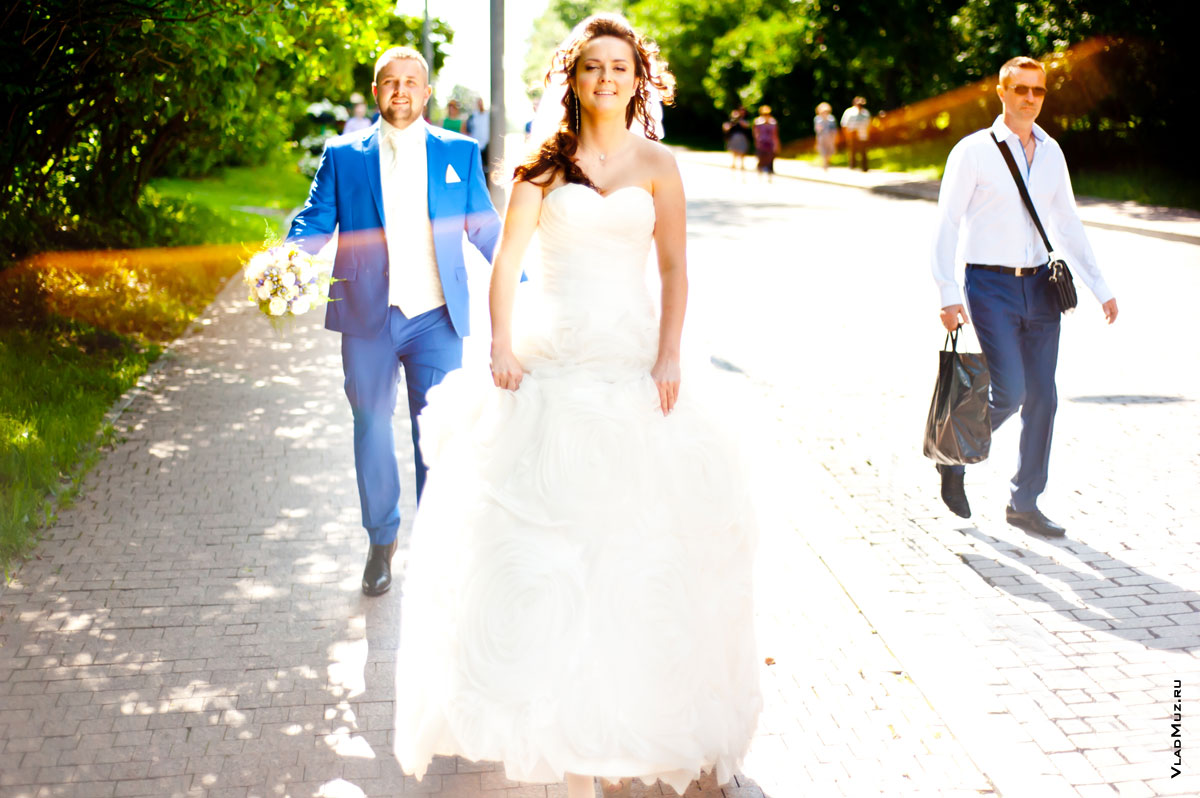 Невеста идет впереди, жених сзади помогает нести шлейф свадебного платья