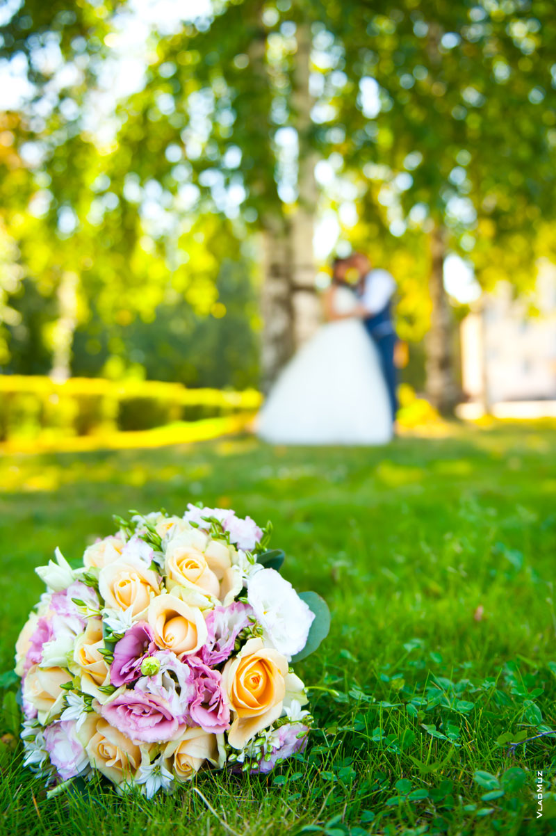 Фото свадебного букета в фокусе на траве, вдали в расфокусе - жених с невестой