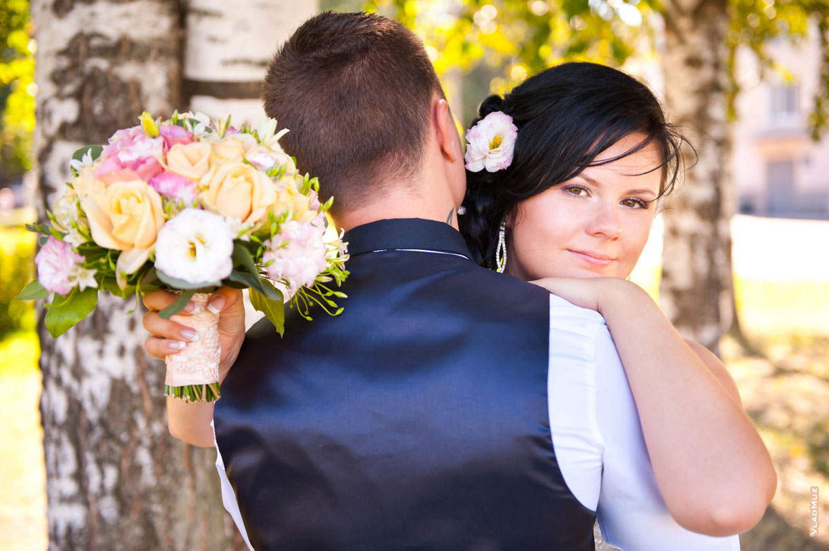 Фотопортрет невесты с букетом в объятиях с женихом (жених стоит спиной, невеста спокойно смотрит в кадр)