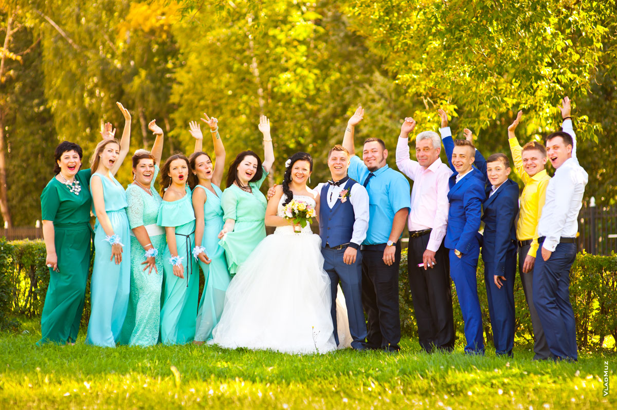 Веселое групповое свадебное фото молодоженов с друзьями на лужайке в парке