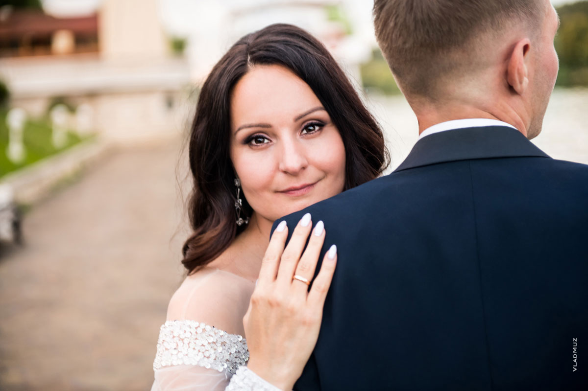 Портретное фото невесты с акцентом на глазах и кольцом на руке, жених стоит спиной