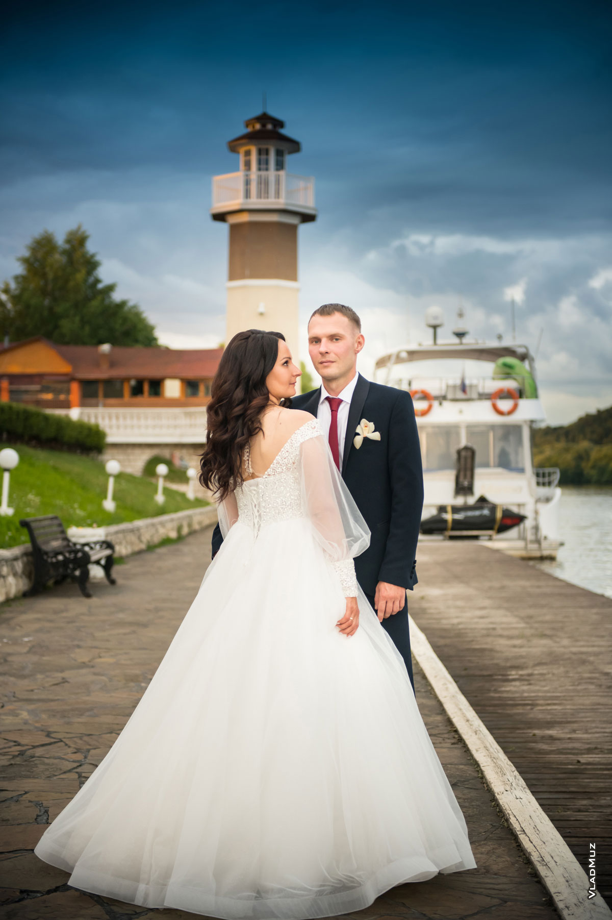 Фото свадебной пары в полный рост на фоне маяка комплекса «Белый берег»: жених смотрит в кадр, невеста — на жениха