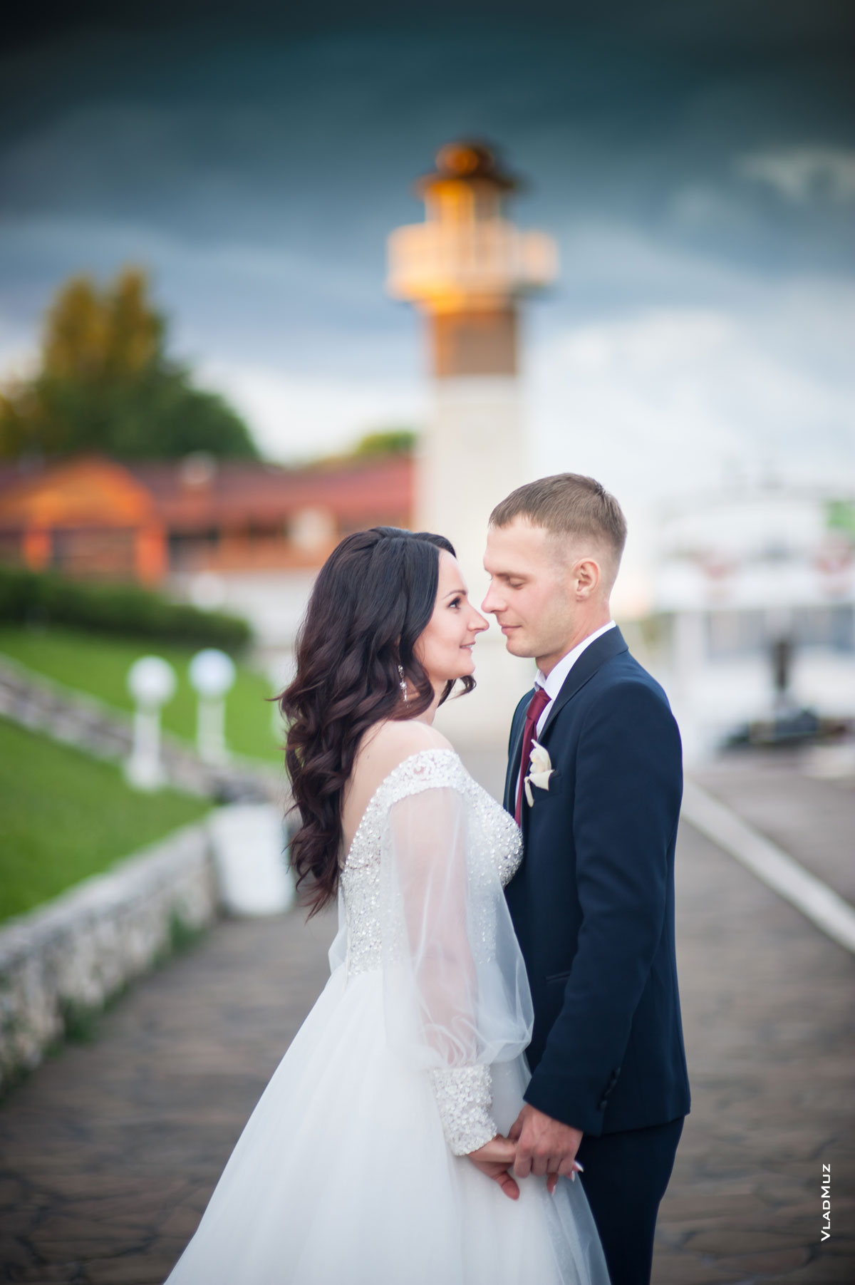 Фото жениха с невестой напротив друг друга, держащихся за руки, на фоне маяка комплекса «Белый берег»