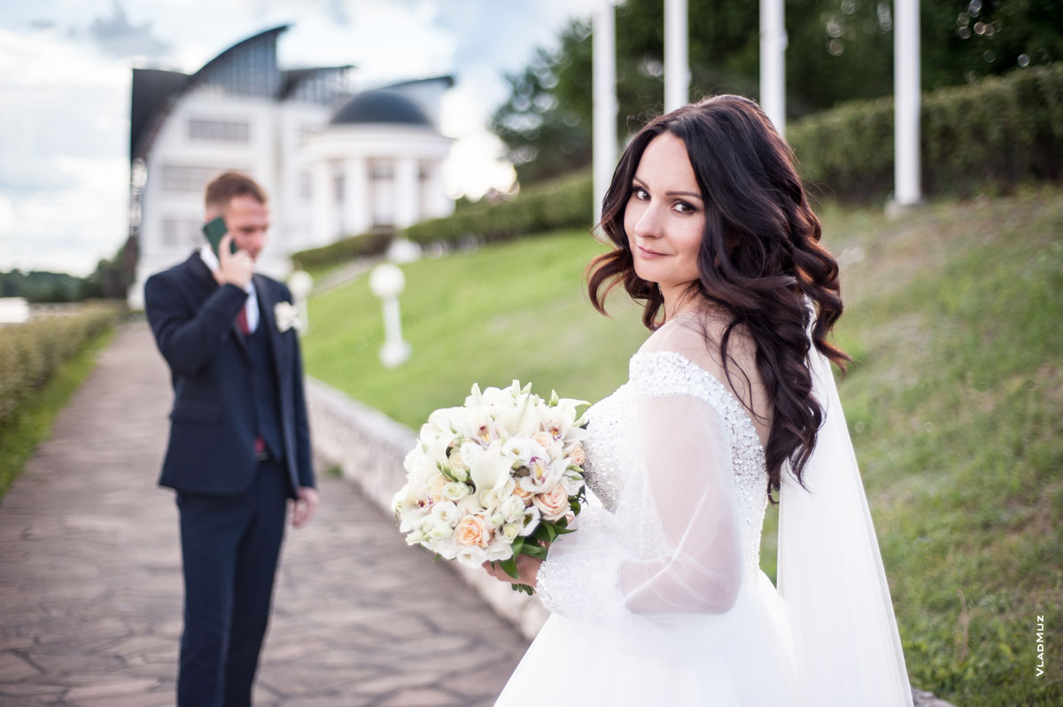 Фото невесты с букетом в фокусе, вдали жених говорит по телефону — в расфокусе