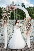 Фото смеющейся невесты с букетом в полный рост в свадебной арке на берегу реки