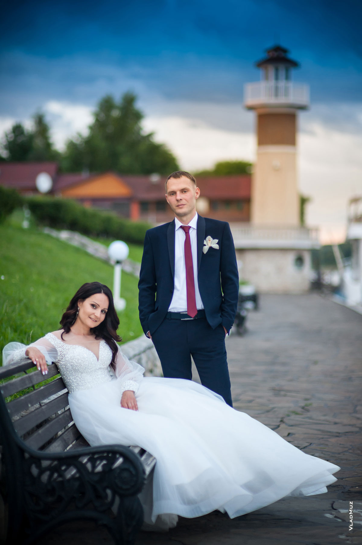 Фото невесты, сидя на лавочке, и жениха в полный рост, стоящего рядом, на фоне маяка