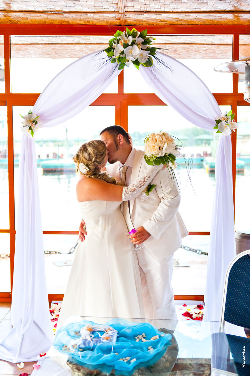Фото поцелуя молодоженов в свадебной арке в ресторане «Малибу» на воде, в Пирогово