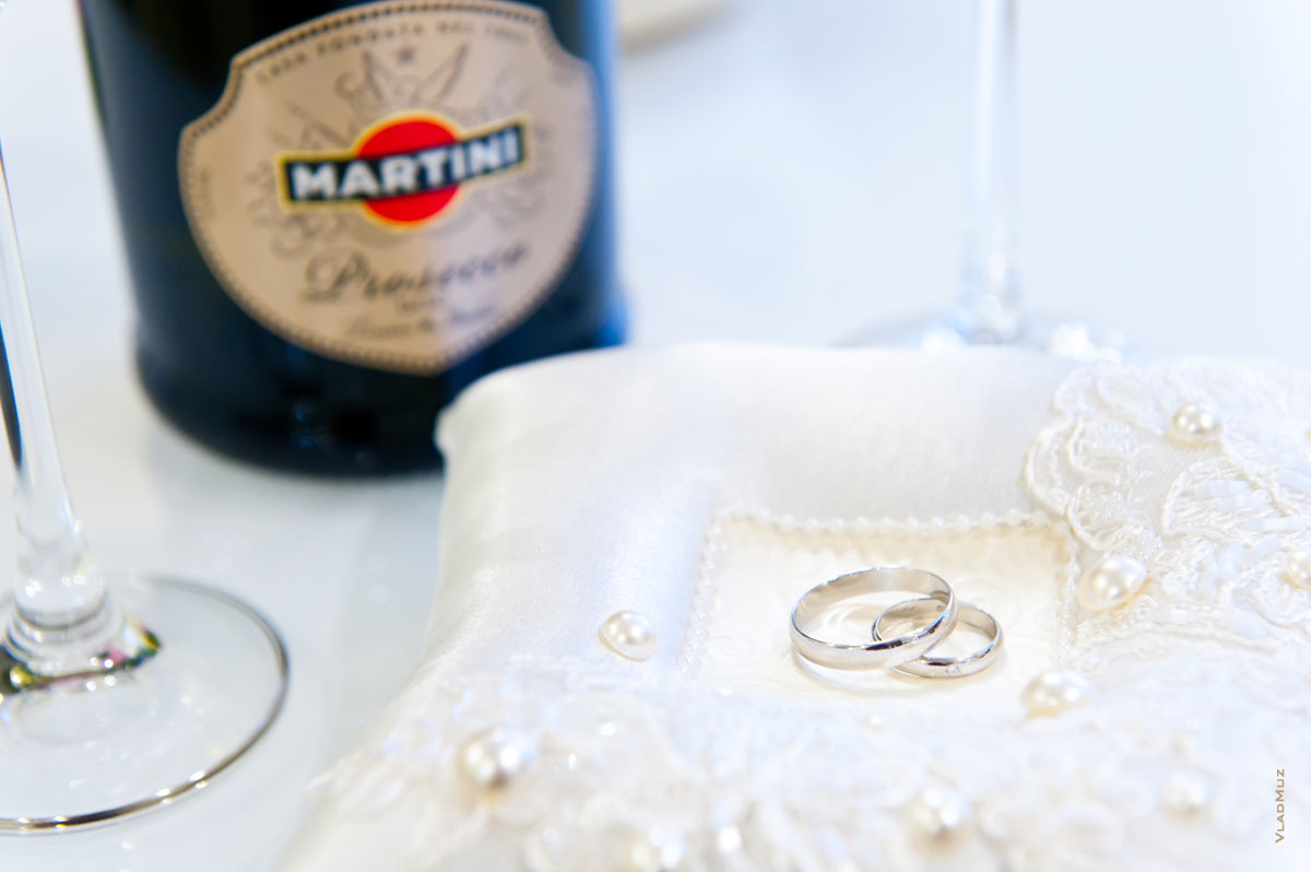 Фото свадебных колец на белой подушке, бутылка Martini в расфокусе на дальнем плане