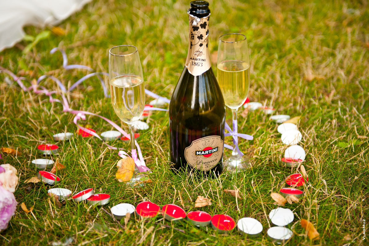 Фото бутылки Martini с бокалами шампанского на траве в окружении свечей в форме сердца