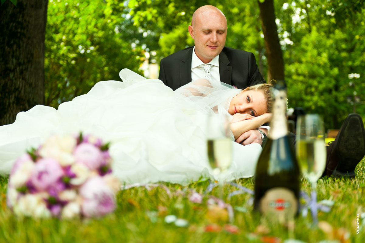 Фото букета невесты и бутылки Martini с бокалами в нерезкости, вдали — жених и невеста на траве