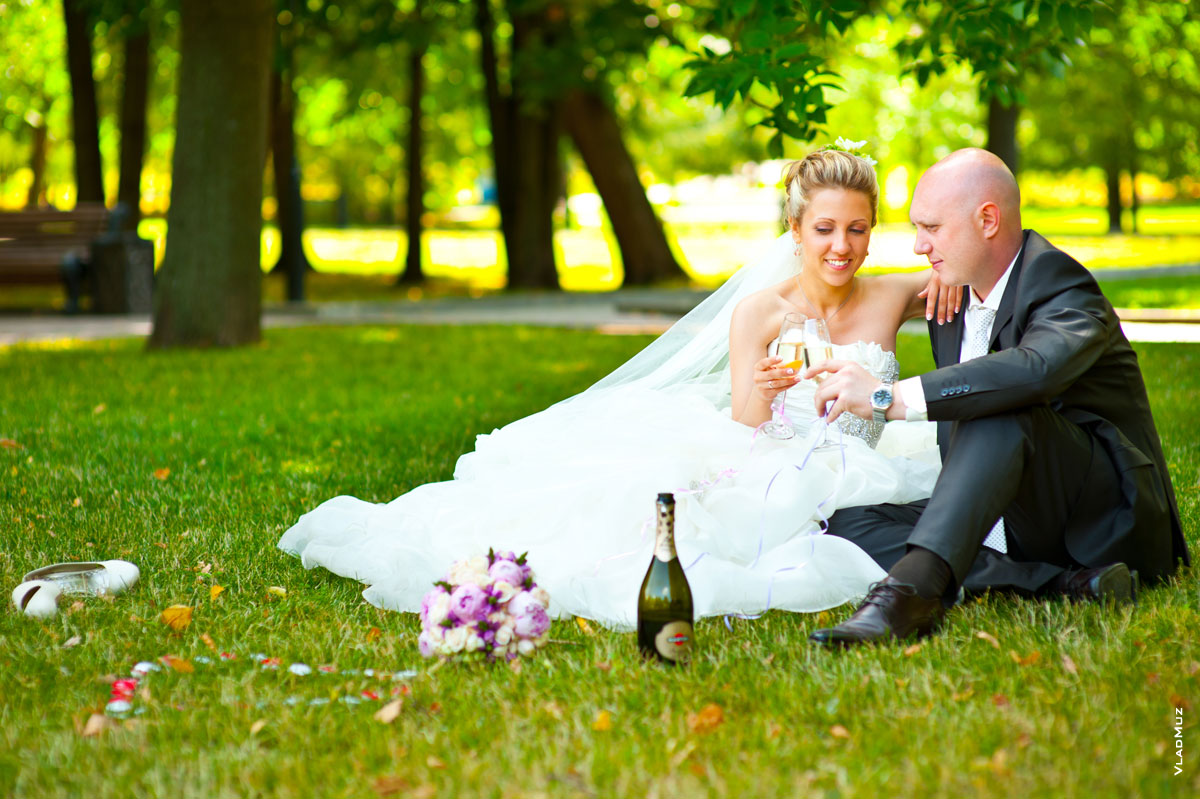 На фото жених и невеста с бокалами шампанского сидят на лужайке в Екатерининском парке Москвы