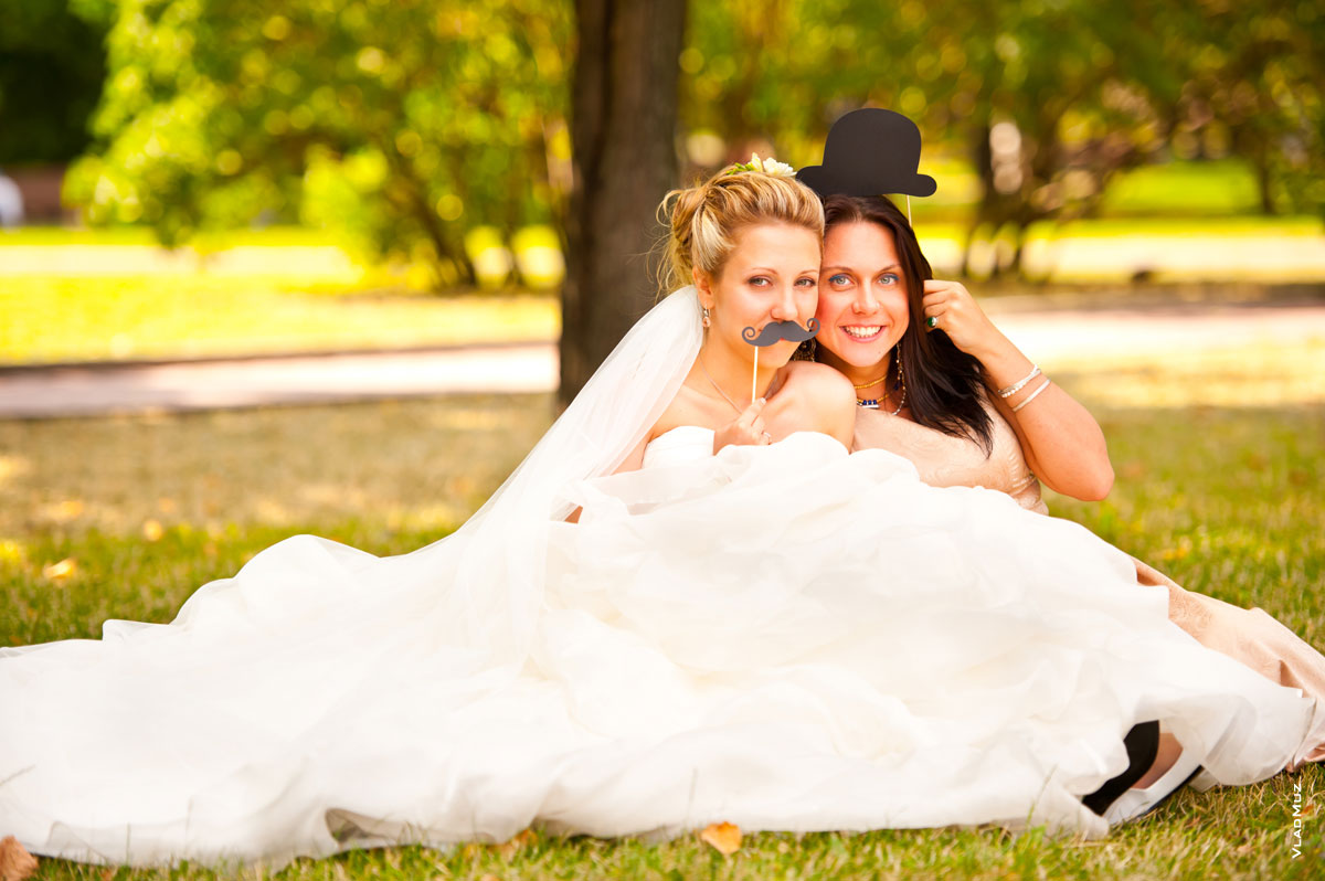 Прикольное фото невесты со своей сестрой с бутафорскими накладными усами и котелком на палочках
