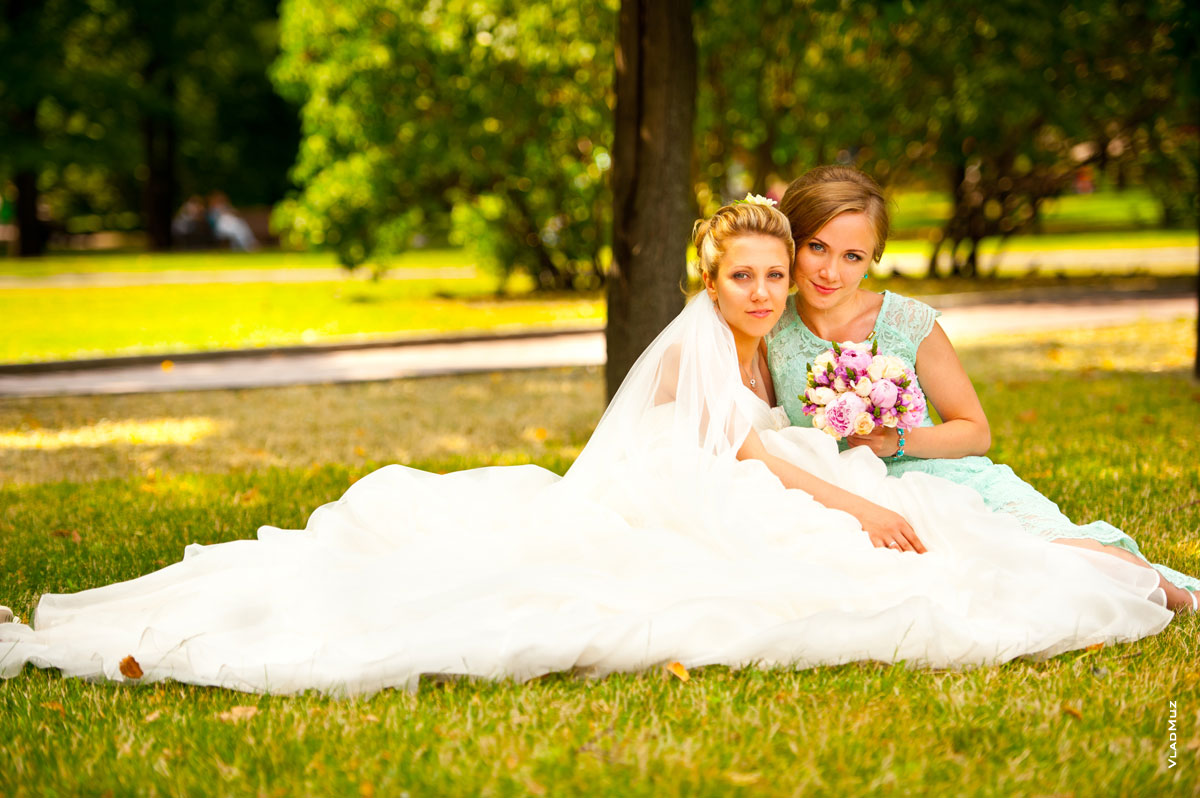 Фото невесты в свадебном платье с большим шлейфом и почетной свидетельницы, сидящих на лужайке в Екатерининском парке