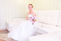 Фото красивой невесты в платье с букетом, сидящей на белом диване