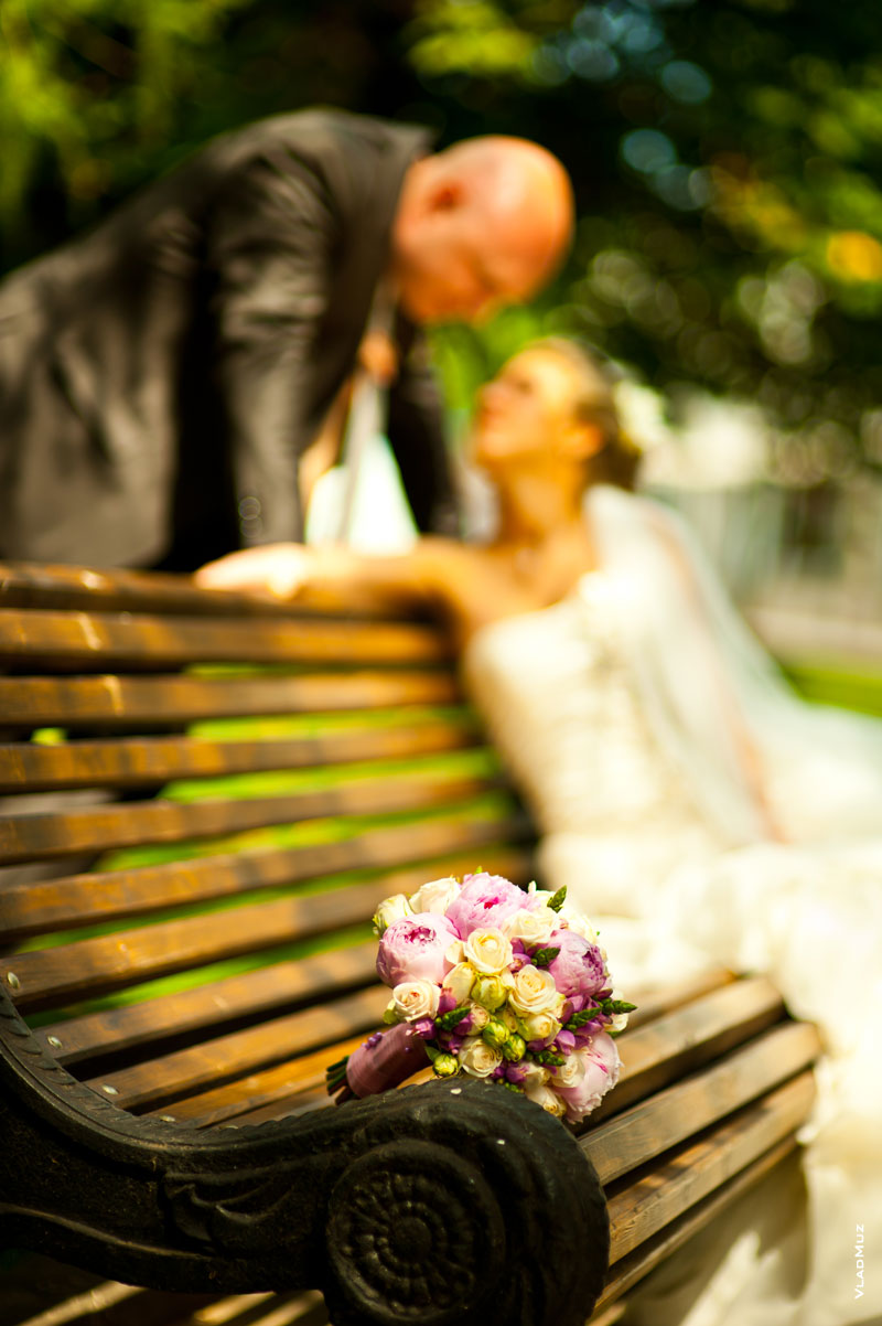 Фото свадебного букета на лавочке в резкости, а жених и невеста вдали — в красивой нерезкости