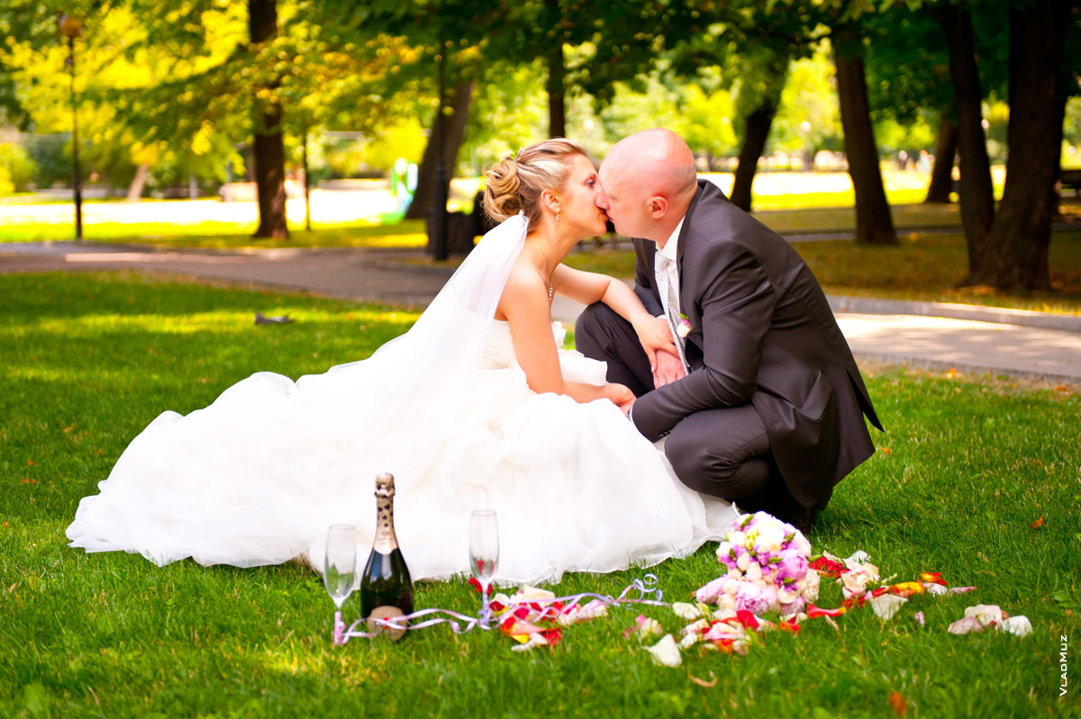 Фото поцелуя жениха и невесты на лужайке в парке. Рядом — бутылка шампанского, бокалы, букет и лепестки роз на зеленой траве