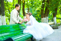 Невеста расположилась на лавочке, жених стоит с другой стороны перед невестой. Здесь делать парные свадебные фотографии легко, потому что в парке просторно