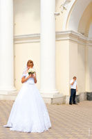 Фото невесты с букетом в полный рост на переднем плане, жених вдали говорит по телефону