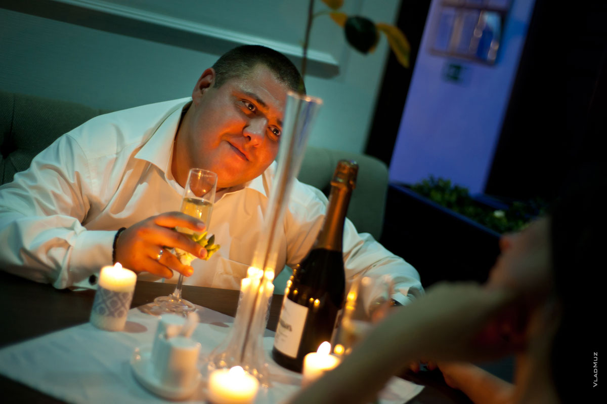 Фото жениха за столом в кафе с бокалом шампанского при свете свечей