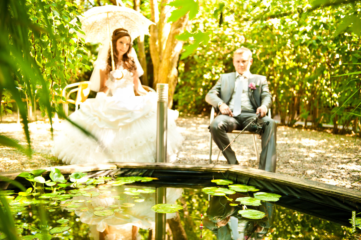 Свадебное фото у пруда: жених ловит рыбу удочкой, невеста с зонтом сидит рядом